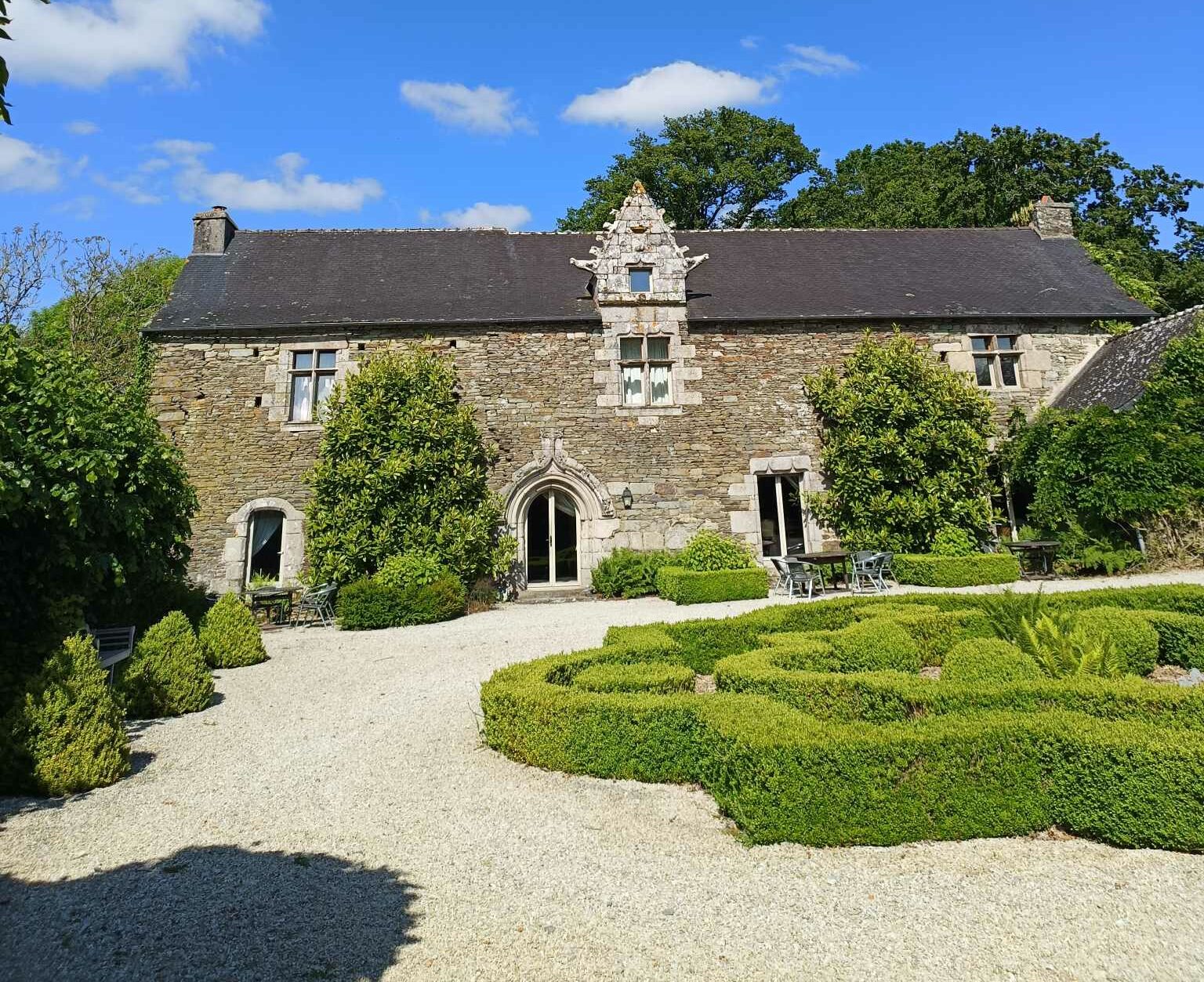 Façade en pierre du Manoir de Kerledan, une bâtisse du XVe siècle entourée de verdure, avec ses volets bleus et son jardin fleuri en premier plan.